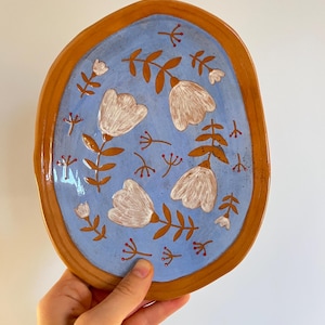 Può includere: Un piatto in ceramica dipinto a mano con interno azzurro e bordo marrone. Il piatto presenta fiori bianchi con steli e foglie marroni e piccoli accenti rossi. Il piatto è tenuto da una mano.