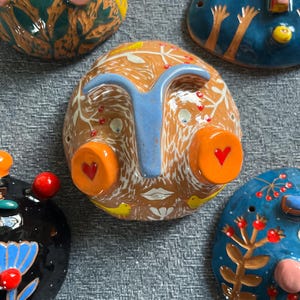 May include: Five ceramic wall hangings with whimsical faces and colorful details. Each piece features unique designs and textures, including flowers, birds, and geometric patterns.