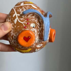 May include: A brown ceramic piggy bank with white and blue painted details. The piggy bank has a heart-shaped nose, a blue ear, and a white eye. The piggy bank is decorated with yellow birds and red berries.