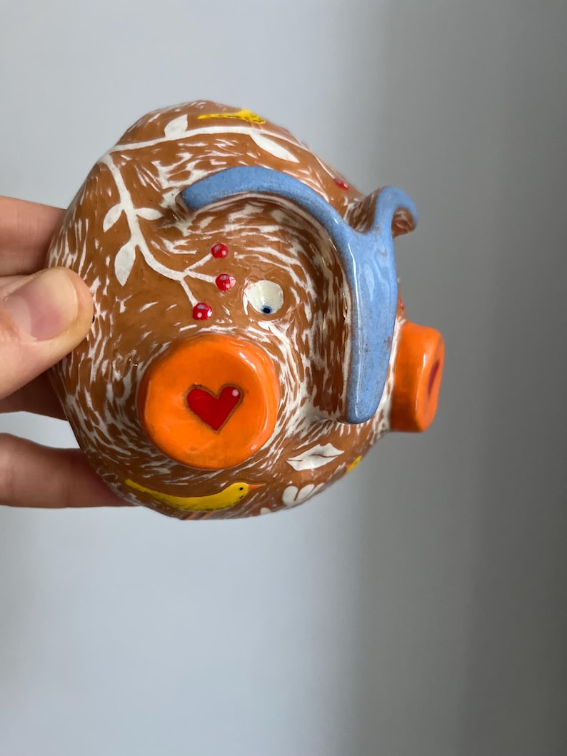 May include: A brown ceramic piggy bank with white and blue painted details. The piggy bank has a heart-shaped nose, a blue ear, and a white eye. The piggy bank is decorated with yellow birds and red berries.