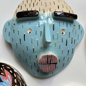 May include: A ceramic face sculpture with a light blue face, black eyebrows, and a light beige head. The face has a nose, two eye holes, and a circular mouth with pink and blue stripes. Black lines and dots decorate the face.