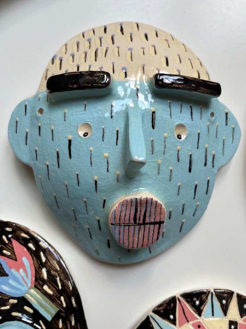 May include: A ceramic face sculpture with a light blue face, black eyebrows, and a light beige head. The face has a nose, two eye holes, and a circular mouth with pink and blue stripes. Black lines and dots decorate the face.