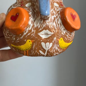 May include: A ceramic face planter with a brown base, white accents, and blue and orange details. The face has two large orange circles with red hearts, a white mouth, and two yellow birds on the bottom.