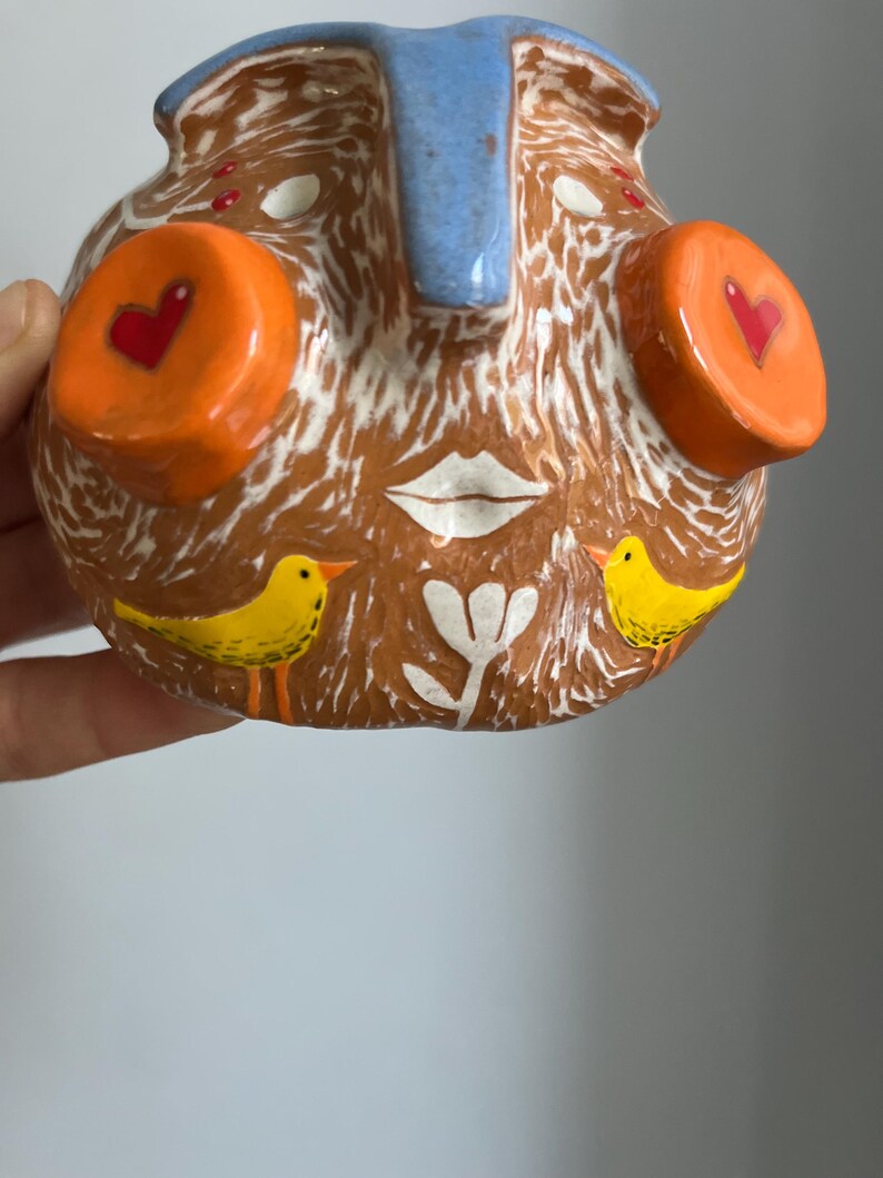 May include: A ceramic face planter with a brown base, white accents, and blue and orange details. The face has two large orange circles with red hearts, a white mouth, and two yellow birds on the bottom.