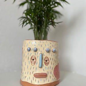 Puede incluir: Un macetero de cerámica con forma de cara con ojos azules, nariz azul y boca rosa. El macetero está decorado con líneas azul claro y blancas y tiene una base marrón claro.