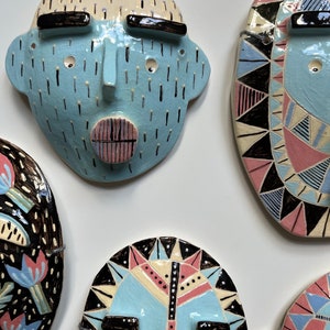 May include: A collection of ceramic face masks in various designs. The masks feature a light blue base color with black, pink, and white accents. Some masks have geometric patterns, while others have a more abstract design. The masks are mounted on a white surface.