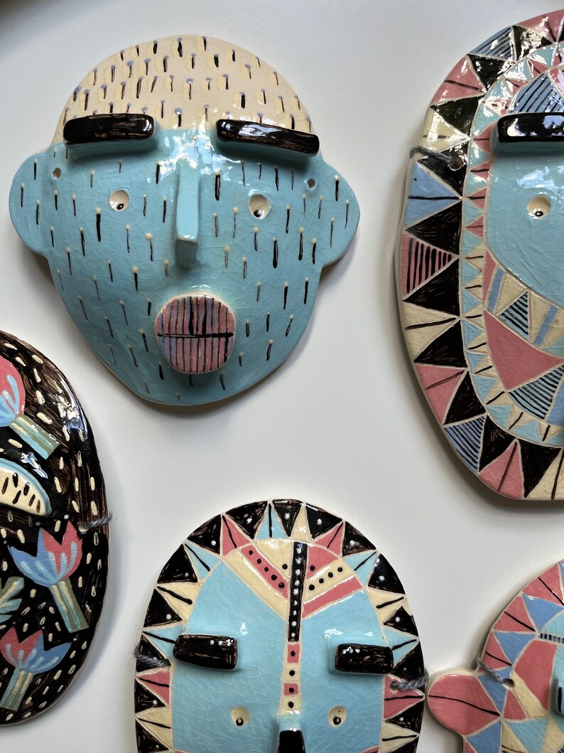 May include: A collection of ceramic face masks in various designs. The masks feature a light blue base color with black, pink, and white accents. Some masks have geometric patterns, while others have a more abstract design. The masks are mounted on a white surface.