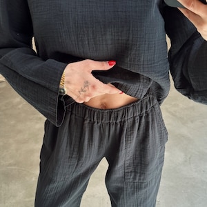 Könnte beinhalten: Ein schwarzes, langärmeliges Hemd und eine passende Hose aus weichem, strukturiertem Stoff. Das Hemd hat eine lockere Passform und die Hose hat einen elastischen Bund. Die Person trägt schwarze Schuhe.
