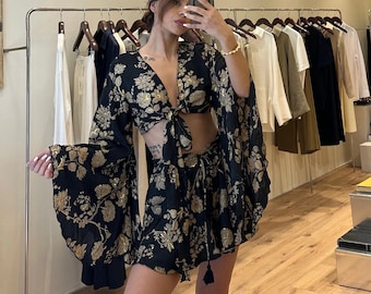 Puede incluir: Conjunto de dos piezas con estampado floral negro con top anudado y shorts. El conjunto presenta detalles florales dorados y mangas acampanadas. Los shorts tienen cintura con cordón y detalle de borla. El conjunto se exhibe en una tienda de ropa.