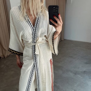 May include: A white cotton robe with black and red embroidery. The robe has a long, flowing silhouette and a tie belt. The robe is worn over a pair of white trousers.