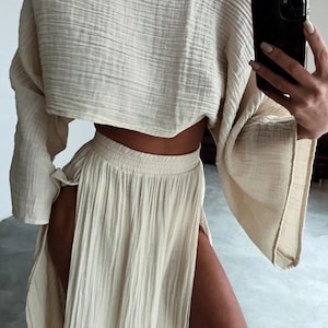 May include: A two-piece cream-colored outfit. The top is a cropped, long-sleeved shirt with a wide neckline. The skirt is a maxi skirt with a high slit on the side. The fabric has a textured, crinkled appearance.