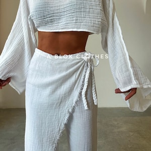 May include: A white two-piece outfit with a cropped, long-sleeved top and a wrap skirt. The top has a relaxed fit and the skirt has a frayed hem. The outfit is made of a lightweight, textured fabric.