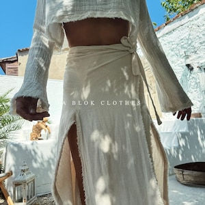 May include: A white linen wrap skirt with a frayed hem and a side slit. The skirt is tied at the waist with a matching fabric belt. The skirt is worn with a white linen top with long sleeves and a frayed hem.