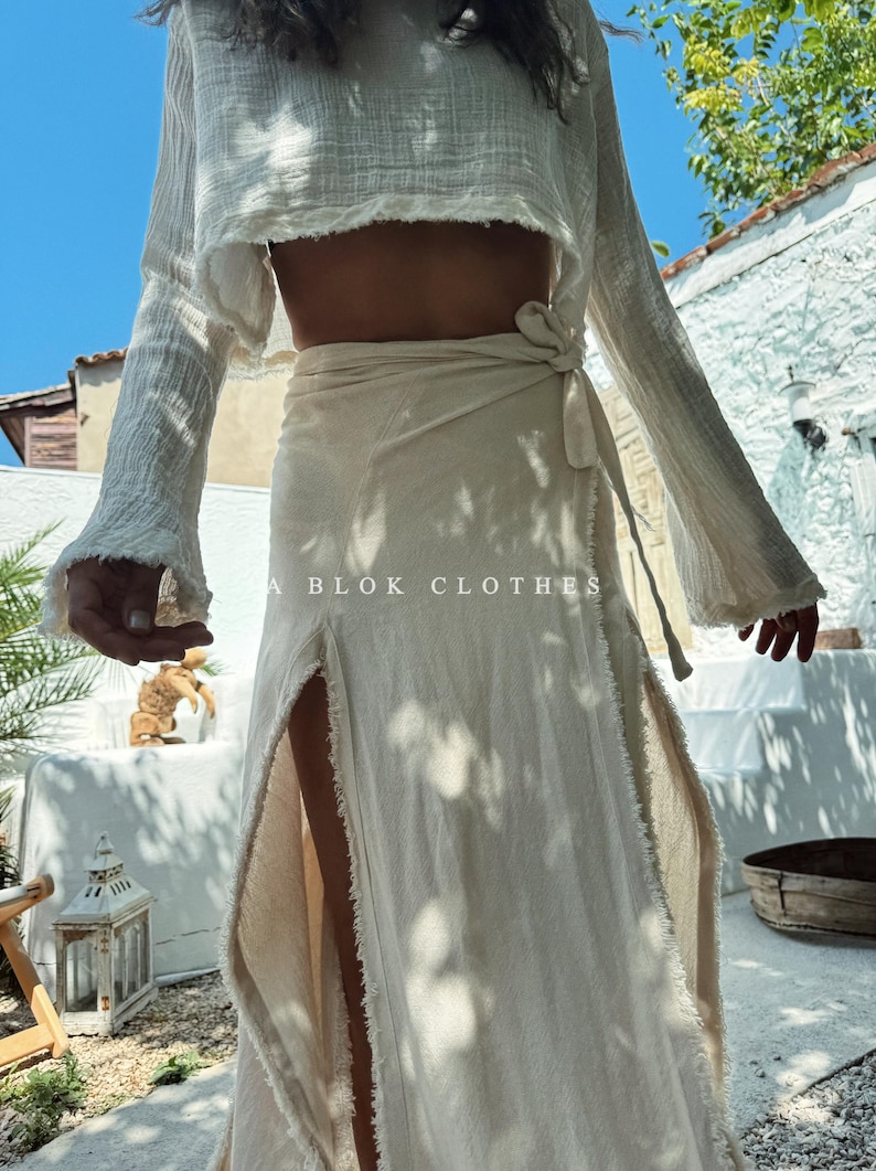 May include: A white linen wrap skirt with a frayed hem and a side slit. The skirt is tied at the waist with a matching fabric belt. The skirt is worn with a white linen top with long sleeves and a frayed hem.