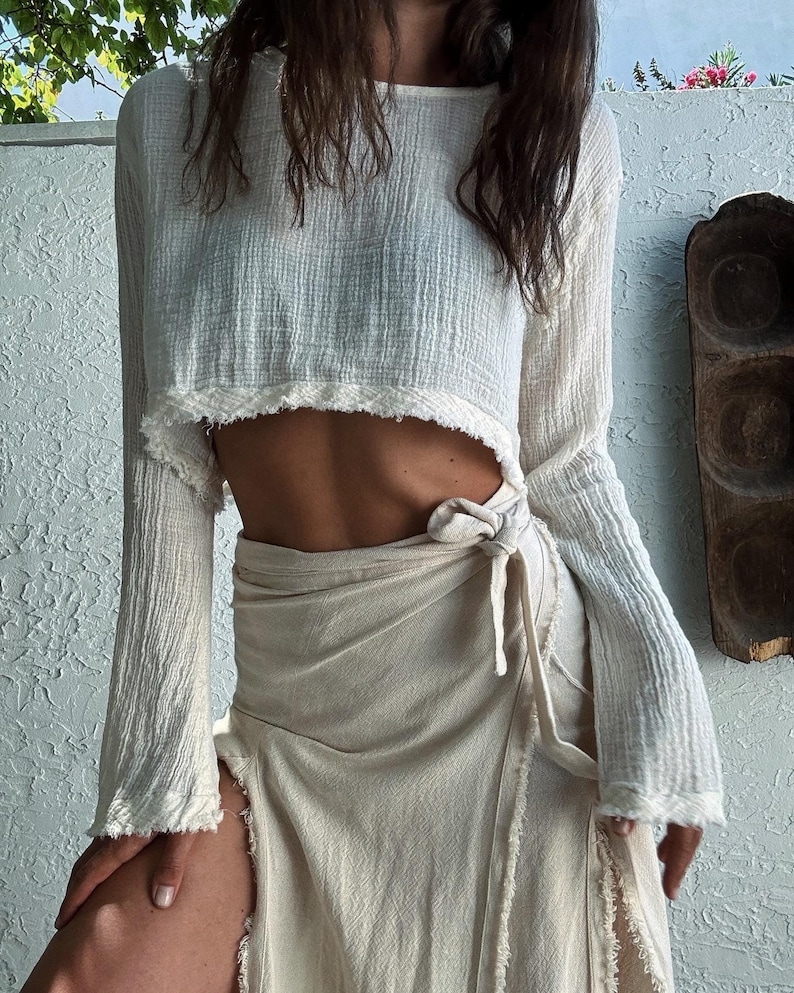 May include: A white, long-sleeved, cropped top with a frayed hem. The top is worn with a cream-colored, wrap skirt that has a frayed hem and a tie at the waist.