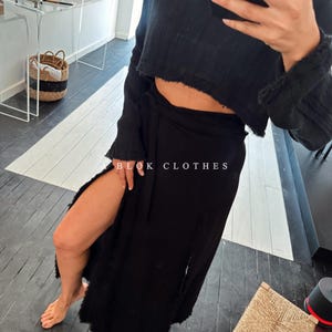 May include: A black wrap skirt with a high slit on the left side. The skirt is tied at the waist with a black sash. The skirt is made of a soft, flowing fabric. The skirt is paired with a black cropped sweater.