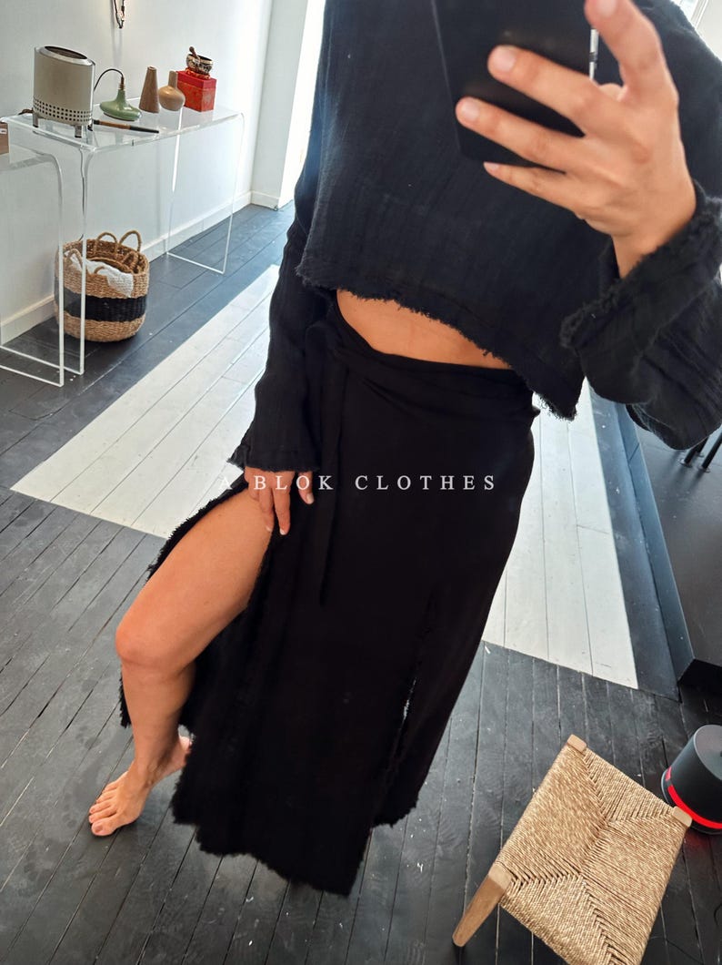 May include: A black wrap skirt with a high slit on the left side. The skirt is tied at the waist with a black sash. The skirt is made of a soft, flowing fabric. The skirt is paired with a black cropped sweater.