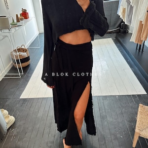 May include: A black, long-sleeved, cropped top with a wrap skirt. The skirt has a side slit and a frayed hem. The model is wearing the outfit in a store with a wooden floor.