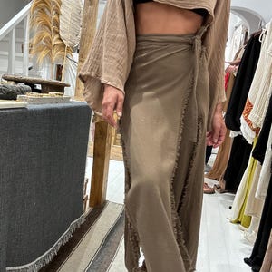 May include: A brown, long, wrap skirt with frayed edges and a matching brown, cropped top with long, wide sleeves. The top is tied at the waist with a brown fabric belt.