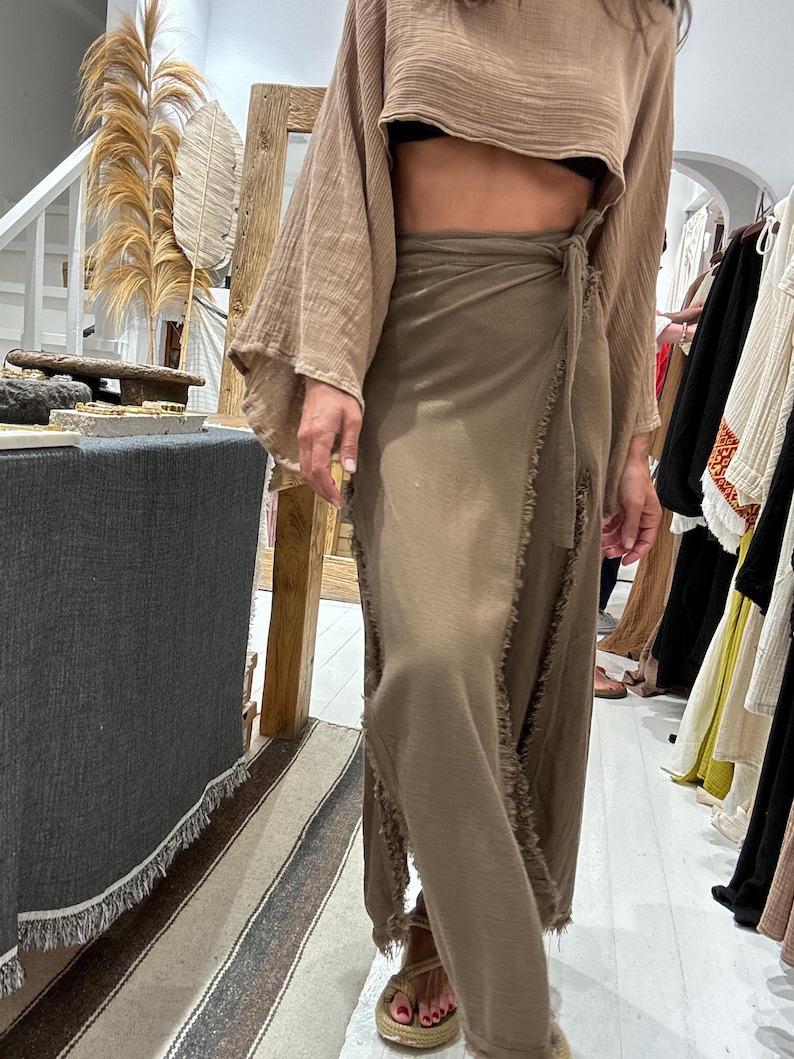 May include: A brown, long, wrap skirt with frayed edges and a matching brown, cropped top with long, wide sleeves. The top is tied at the waist with a brown fabric belt.