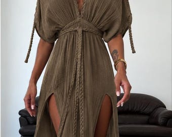 Puede incluir: Un vestido largo de color marrón oliva con un escote en V profundo, mangas cortas con detalles de lazos trenzados y un cinturón trenzado en la cintura. La prenda está confeccionada en tela texturizada y tiene dos aberturas laterales altas.