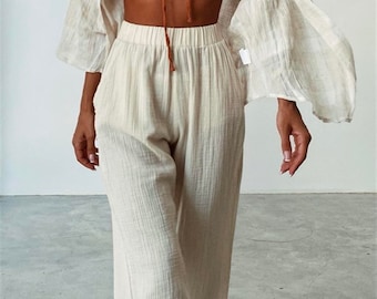 May include: A rust-colored strapless tube top with a center tie detail, paired with high-waisted, wide-leg cream-colored trousers and a matching cream-colored open-front long-sleeve shirt with a textured, crinkled fabric finish.