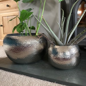May include: Two silver hammered metal planters with a green leafy plant in one and a green aloe plant in the other. The planters are sitting on a black slate surface.
