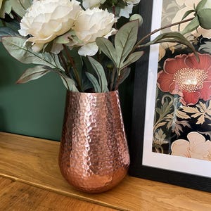 May include: A copper-toned vase with a hammered finish displays white peony flowers and green foliage. The vase is placed on a wooden surface, with a framed floral print visible in the background.
