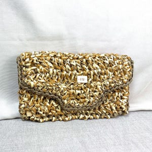 Könnte beinhalten: Eine gewebte Clutch mit Klappenverschluss. Die Tasche besteht aus Naturfasern in Beige- und Brauntönen. Ein kleines rechteckiges Etikett mit den Initialen "IM" befindet sich mittig auf der Vorderseite.
