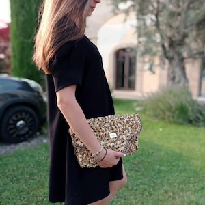 Op de afbeelding: Een zwarte jurk met korte mouwen. Een geweven clutch met klepsluiting wordt voor de jurk gehouden. De clutch is gemaakt van natuurlijk gekleurde vezels en heeft een kleine zilverkleurige metalen plaat.