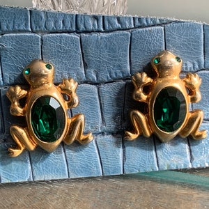 May include: A pair of gold frog earrings with green gemstone accents. The earrings are on a blue background.