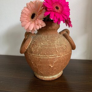 Puede incluir: Un jarrón de terracota con dos asas, decorado con una textura rugosa, que contiene margaritas gerbera rosas y magenta.