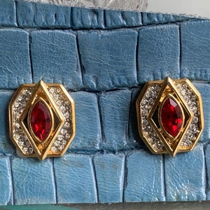 Puede incluir: Un par de pendientes de tono dorado con una piedra roja rodeada de brillantes transparentes.