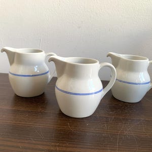 Könnte beinhalten: Drei weiße Keramik-Milchkrüge mit blauem Besatz. Die Krüge haben einen runden Körper und einen kleinen Ausguss.