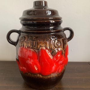 Puede incluir: Un tarro de cerámica marrón con tapa, decorado con pimientos rojos y la palabra "Rumtopf".