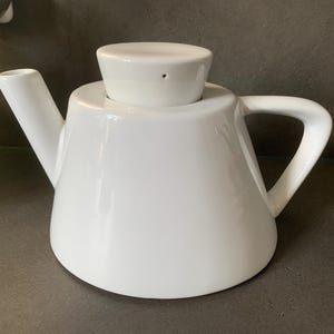 Op de afbeelding: Een witte theepot van keramiek met een conische vorm en een deksel. De theepot heeft een handvat aan de zijkant en een tuit aan de voorkant.