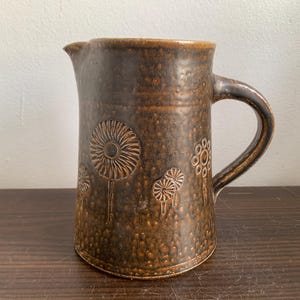 Può includere: Un'anfora in ceramica marrone con una glassa macchiata e un disegno di fiori stilizzati. L'anfora ha un manico e una bocca larga.