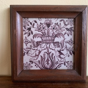 May include: Square framed tile with a floral and cherub design in shades of purple and white. The tile features a central basket of flowers held by two winged cherubs. The frame is made of dark brown wood.