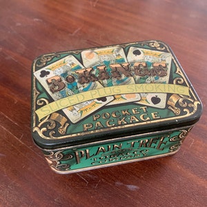May include: Vintage green and gold tin box with a design of playing cards and the text "3 KINGS SLICED PLUG SMOKING POCKET PACKAGE PLAIN TREE TOBACCO COMPANY".