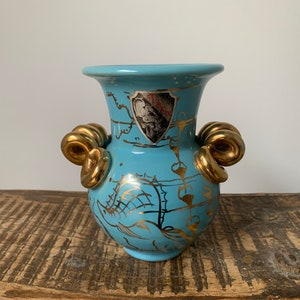 May include: A light blue ceramic vase with gold accents and a decorative design. The vase has two gold handles that are shaped like scrolls.
