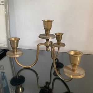 Può includere: Un candelabro in ottone con quattro portacandele. Il candelabro ha un design curvo con un supporto centrale e quattro bracci che si estendono verso l'esterno. I portacandele sono circolari e leggermente svasati. Il candelabro è in ottone e ha una finitura lucida.