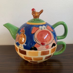 Può includere: Una teiera in ceramica con manico e beccuccio verde. La teiera è decorata con un maiale, un pollo e un motivo a mattoni. Il coperchio della teiera ha un piccolo pollo in cima e la parola "Oink" scritta su di esso.