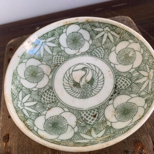 May include: A white ceramic dish with a green floral design and a central image of a crane with outstretched wings. The dish has a scalloped edge and is approximately 5 inches in diameter.