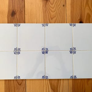 May include: A rectangular arrangement of ten white ceramic tiles with a glossy finish. Each tile features a blue floral design in the corners and center. The tiles are arranged on a wooden surface.