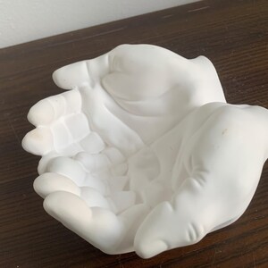 May include: White ceramic sculpture of two hands cupped together, forming a bowl shape.