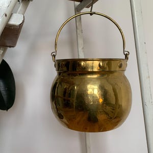 Könnte beinhalten: Ein goldfarbener Metalleimer mit gehämmerter Oberfläche und einem Henkel. Der Eimer hängt an einer weißen Holzleiter.