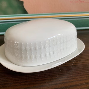 Könnte beinhalten: Weiße Keramik-Butterdose mit einem erhabenen geometrischen Muster. Die Dose hat einen Deckel und einen Boden.