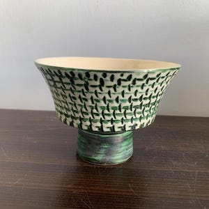 Può includere: Una ciotola in ceramica bianca con un motivo geometrico verde e bianco. La ciotola è su una base a piedistallo.
