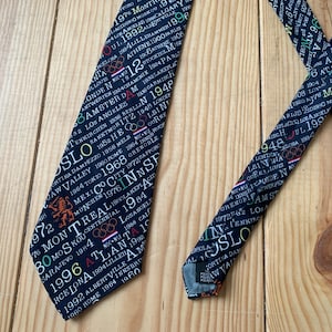 May include: A dark blue necktie with a white print of the Olympic Games host cities and years. The tie features the Olympic rings and the years from 1900 to 1996.