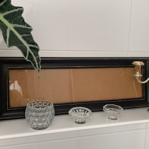 May include: A black picture frame with a brown cardboard insert sits on a white shelf with three clear glass candle holders and a silver candlestick.